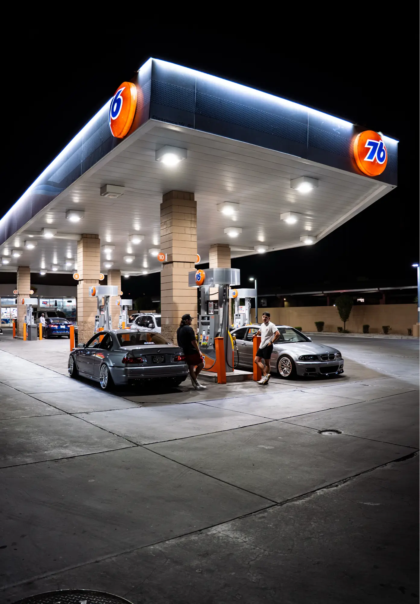 Wide-angle shot of a pair of silver BMW E46 M3s parked together at a gas station at night.