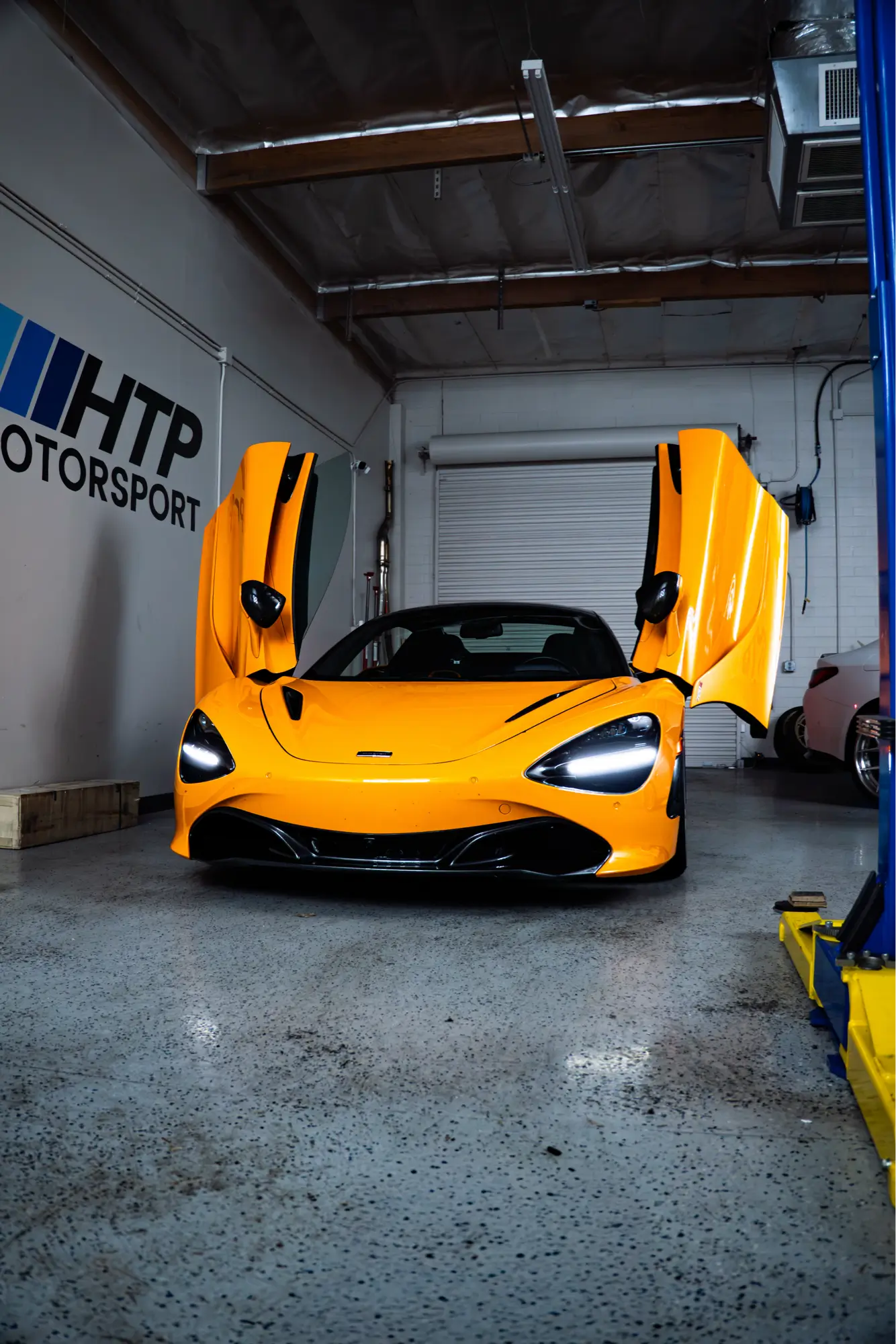 Orange McLaren 720S with dihedral doors raised at the HTP Motorsport shop, the kind of vehicle HTP Motorsport regularly inspects before purchase.
